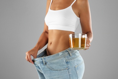 Photo of Young woman in old big jeans with cup of tea showing her diet results on grey background, closeup