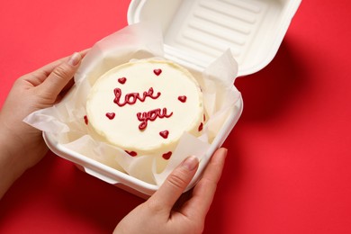 Photo of Woman holding takeaway box with bento cake at red table, closeup. St. Valentine's day surprise