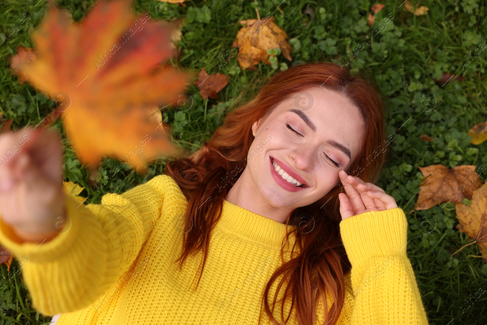Photo of Smiling woman lying on grass and showing autumn leaf, top view