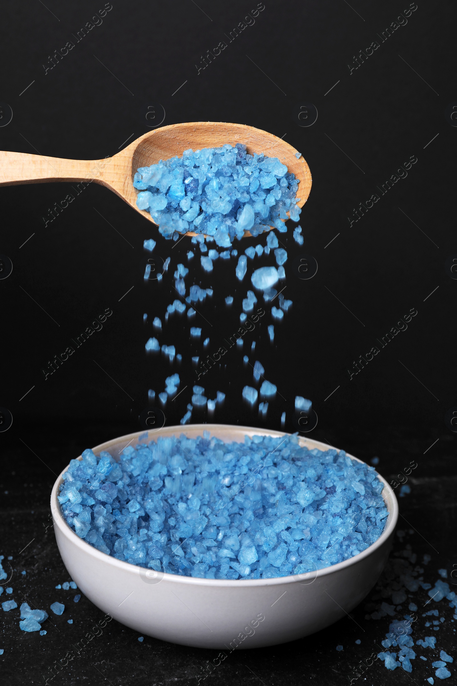 Photo of Pouring blue sea salt from wooden spoon into bowl against black background, closeup