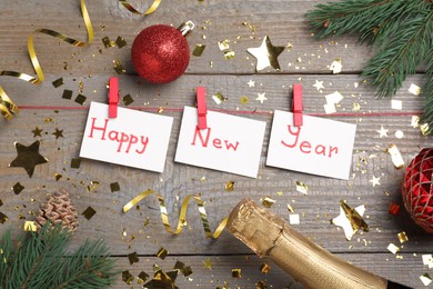 Photo of Phrase Happy New Year made with sheets of paper, sparkling wine and festive decor on wooden background, flat lay