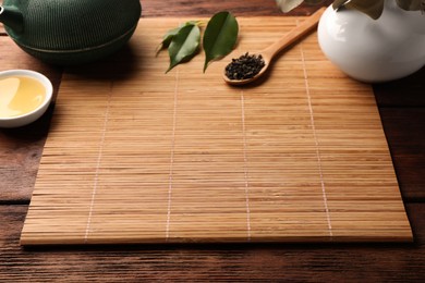 Photo of Bamboo mat, teapot, cup and spoon of dry tea leaves on wooden table, space for text