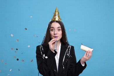 Woman in party hat with blower, piece of tasty cake and flying confetti on light blue background