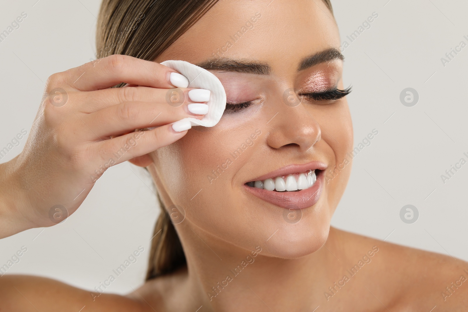 Photo of Beautiful woman removing makeup with cotton pad on light grey background, closeup