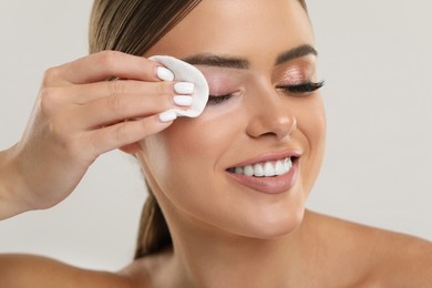 Beautiful woman removing makeup with cotton pad on light grey background, closeup