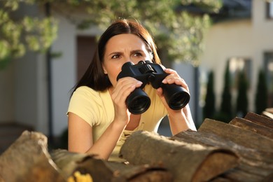 Photo of Concept of private life. Curious young woman with binoculars spying on neighbours over firewood outdoors