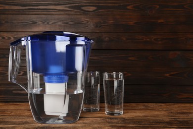 Photo of Filter jug and glasses with purified water on wooden table. Space for text