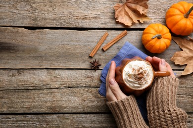 Photo of Woman holding mug of pumpkin spice latte with whipped cream at wooden table, top view. Space for text