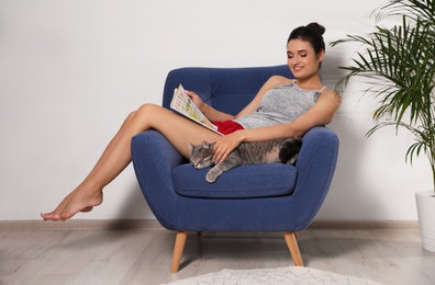 Young woman with cat and magazine on armchair at home. Cute pet