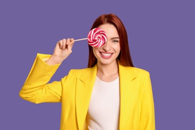 Photo of Stylish redhead woman covering eye with lollipop on purple background