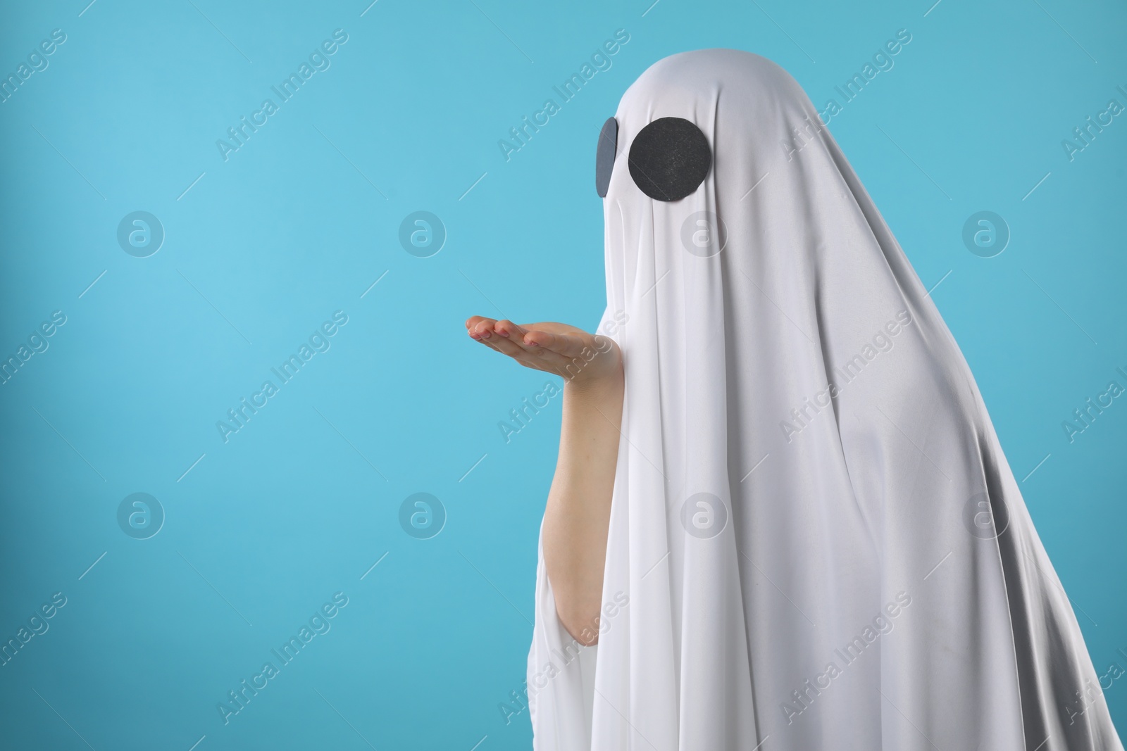Photo of Creepy ghost. Woman covered with white sheet blowing kiss on light blue background
