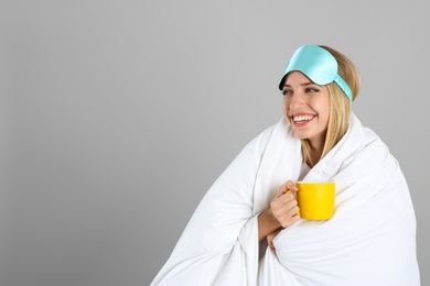 Young woman in sleeping mask wrapped with blanket holding cup on grey background. Space for text