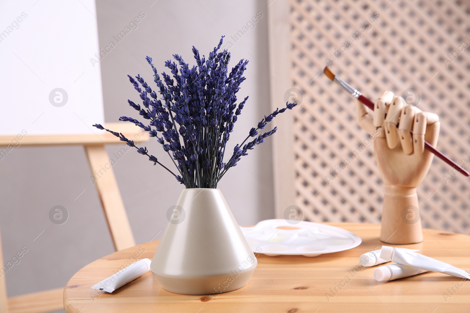 Photo of Bouquet of beautiful preserved lavender flowers, palette, paints and manikin hand with brush on wooden table near easel indoors, closeup