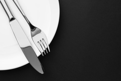 Clean plate, fork and knife on black table, top view. Space for text