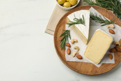 Plate with pieces of tasty camembert cheese, nuts, olives and rosemary on white marble table, flat lay. Space for text