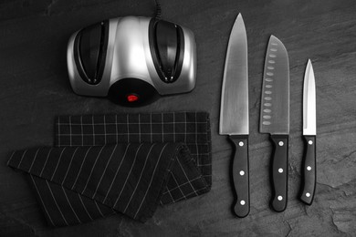 Photo of Modern electrical sharpener and set of knives on black table, flat lay