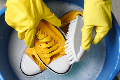 Photo of Woman with gloves and brush cleaning stylish sneakers in wash basin, top view