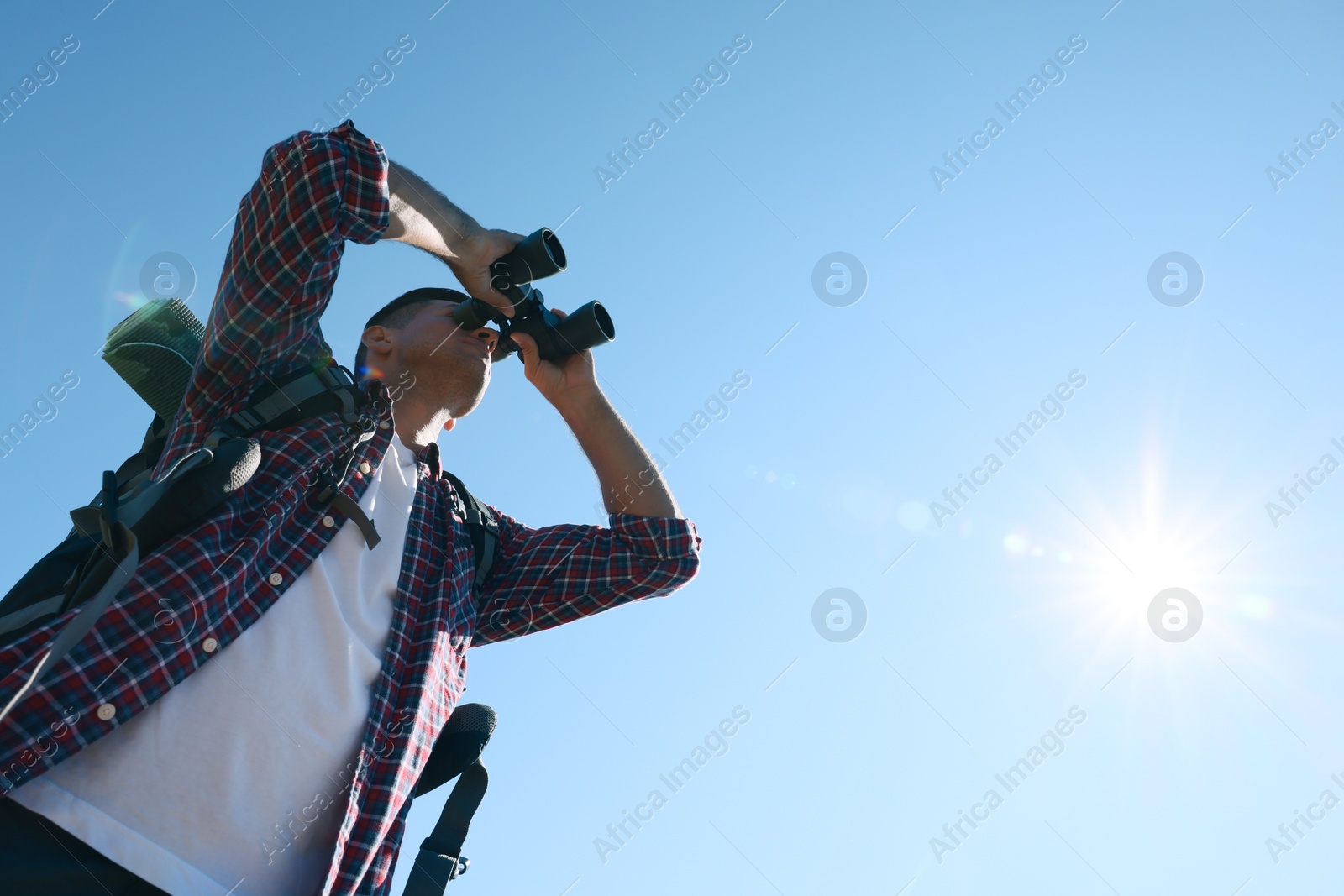 Photo of Tourist with hiking equipment looking through binoculars outdoors on sunny day, low angle view