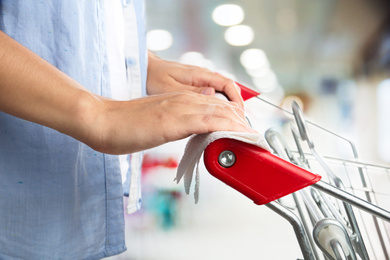Woman holding shopping cart handle with tissue paper at supermarket, closeup. Preventive measure in public places during coronavirus outbreak