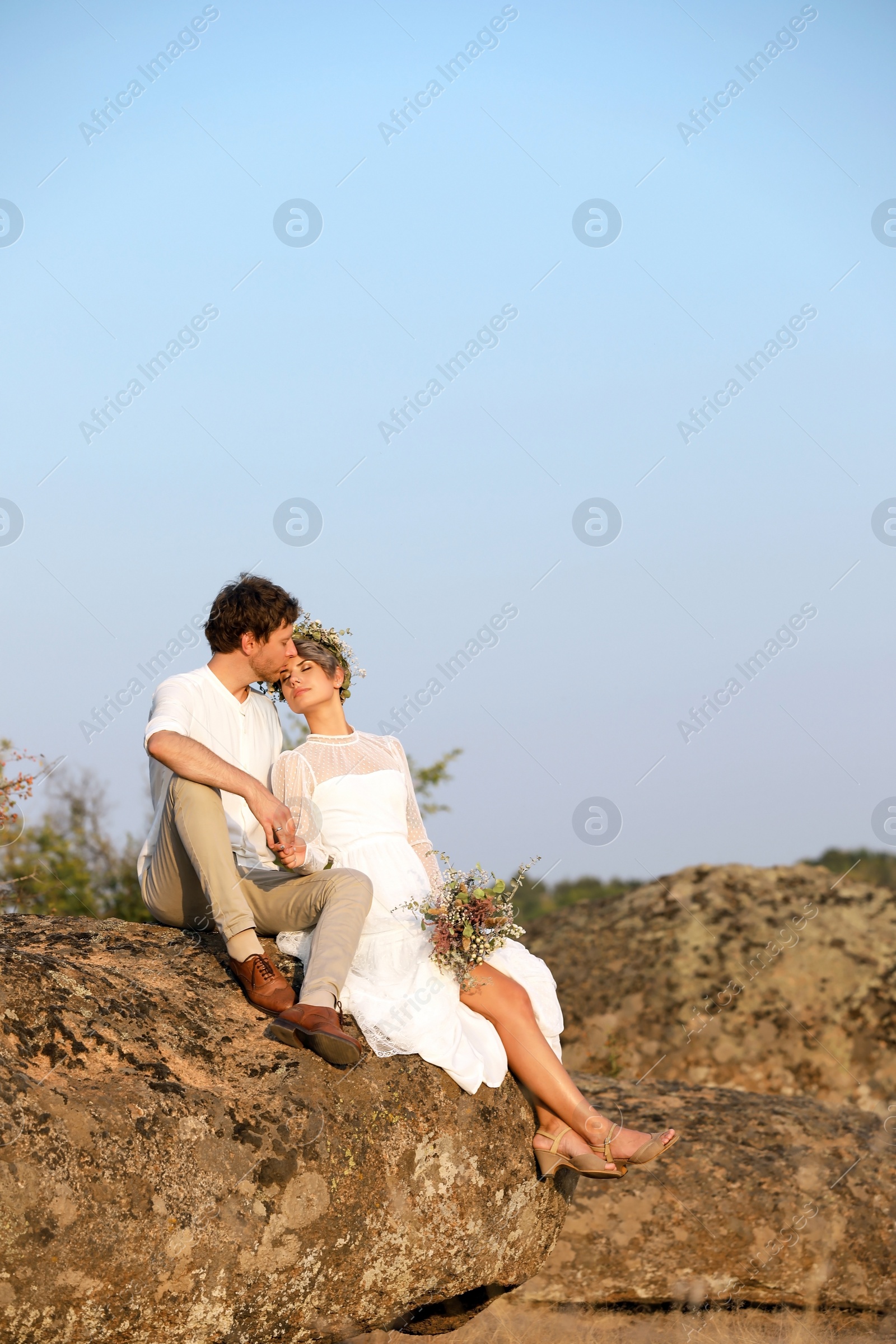 Photo of Happy newlyweds with beautiful field bouquet sitting on rock outdoors