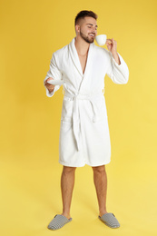 Young man in bathrobe with cup of coffee on yellow background