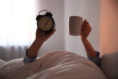 Woman with cup and alarm clock lying in bed, closeup. Morning time