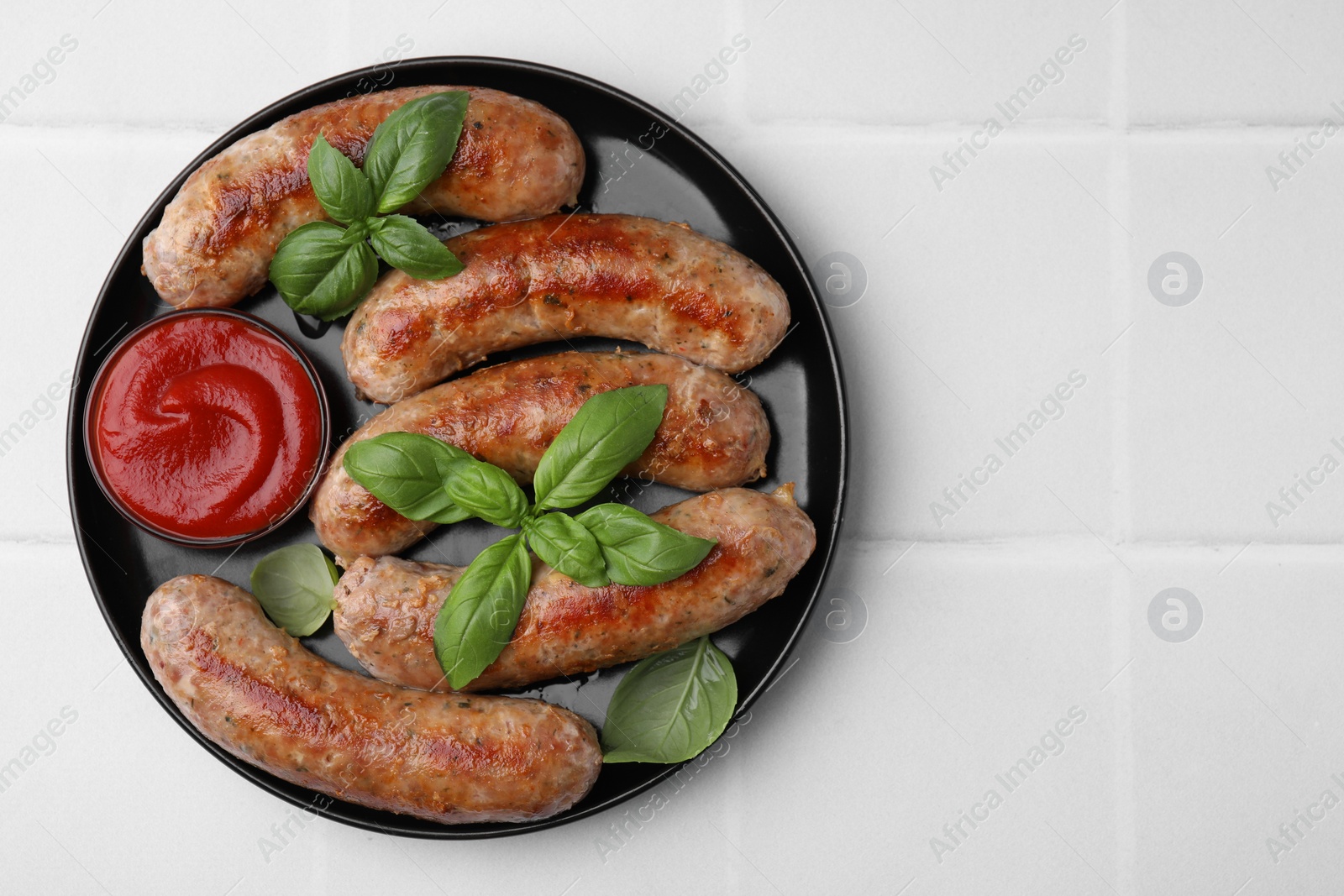 Photo of Plate with tasty homemade sausages, ketchup and basil leaves on white tiled table, top view. Space for text