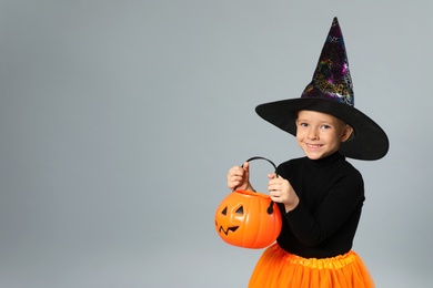 Photo of Cute little girl with pumpkin candy bucket wearing Halloween costume on grey background, space for text