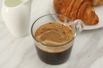 Photo of Aromatic coffee in glass cup, fresh croissant and pitcher on white marble table, closeup