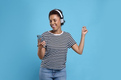 Photo of Happy young woman in headphones dancing with smartphone on light blue background