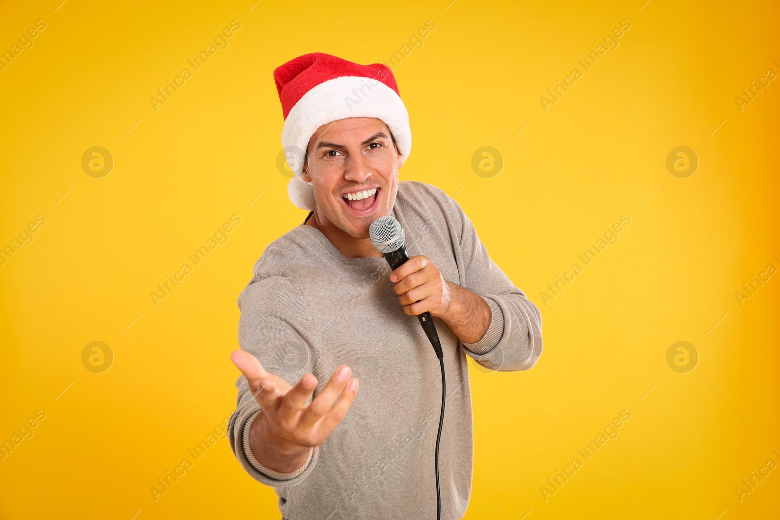 Photo of Happy man in Santa Claus hat singing with microphone on yellow background. Christmas music