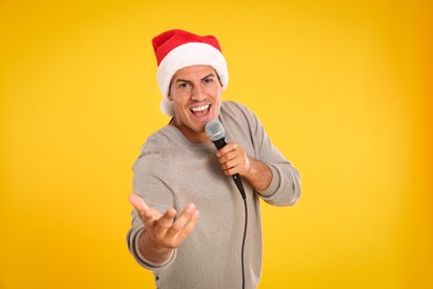 Happy man in Santa Claus hat singing with microphone on yellow background. Christmas music