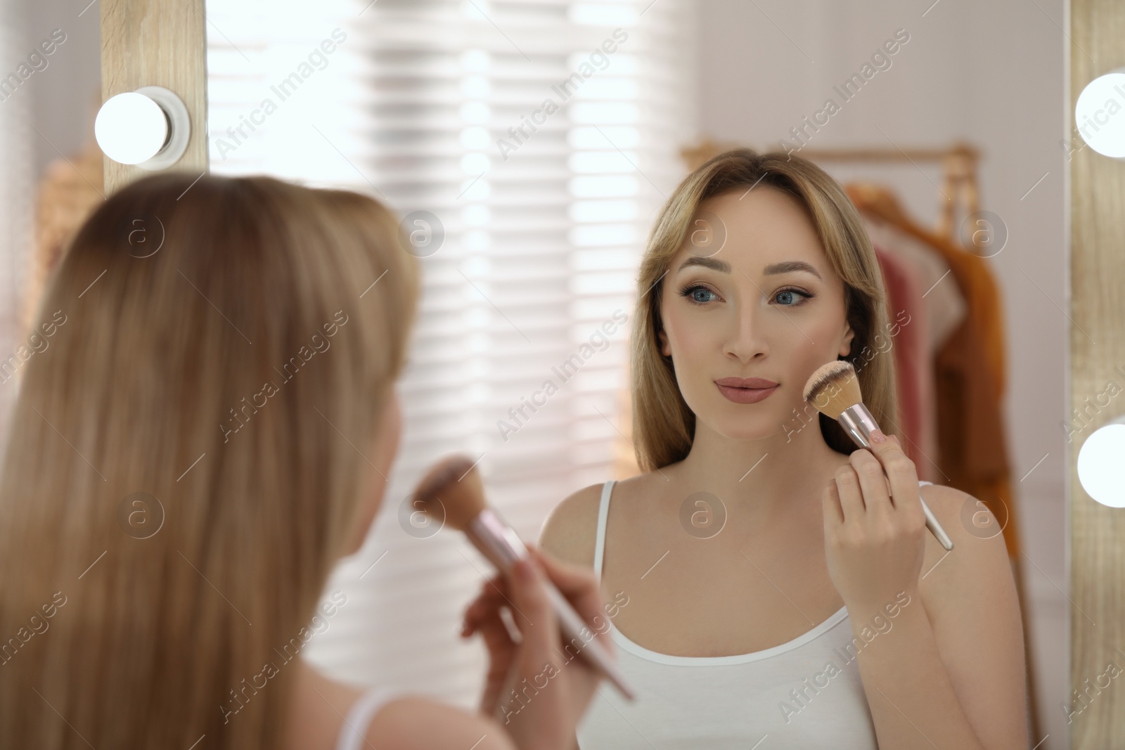 Photo of Beautiful young woman applying face powder with brush in front of mirror at home