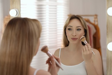Photo of Beautiful young woman applying face powder with brush in front of mirror at home