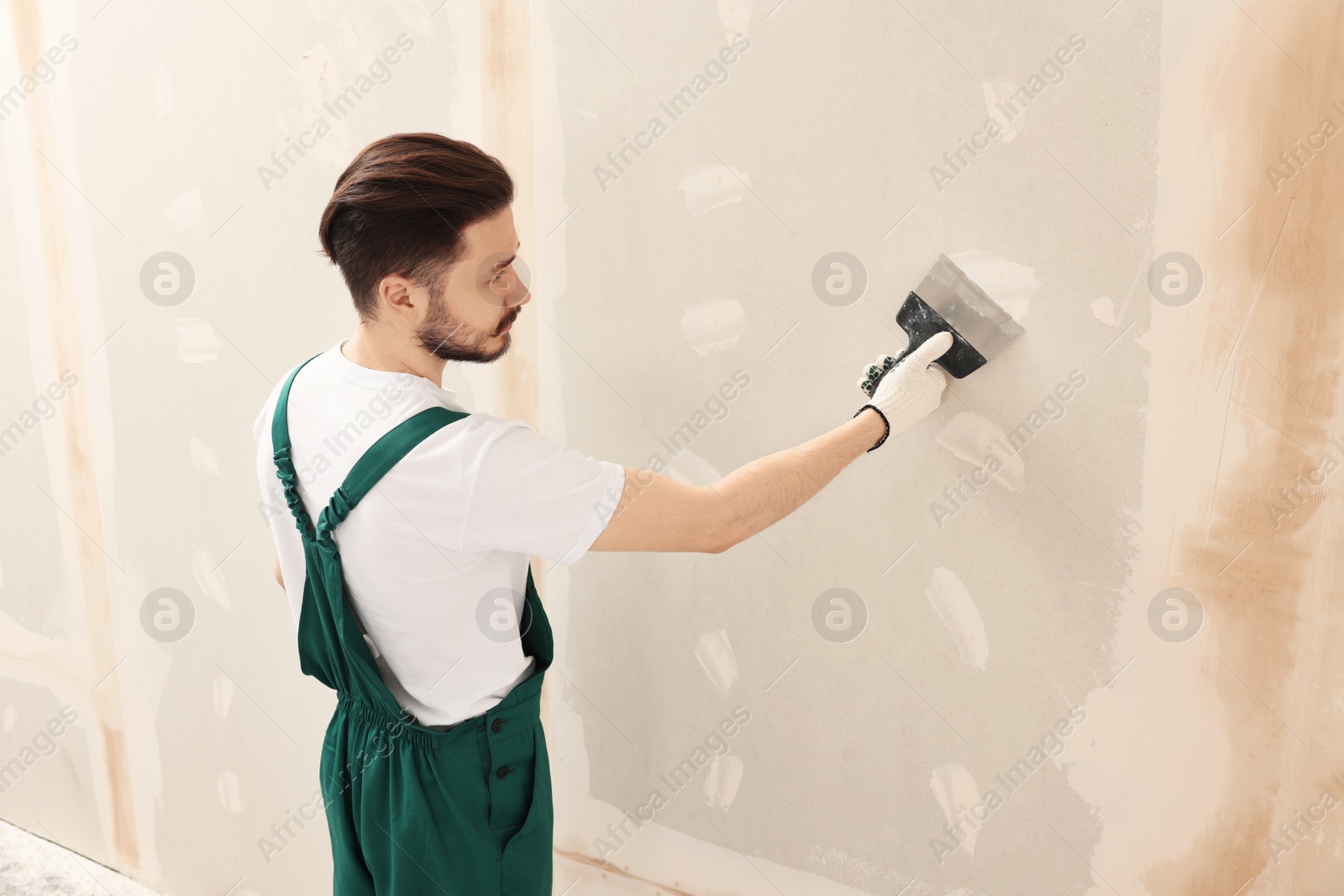 Photo of Worker in uniform plastering wall with putty knife indoors