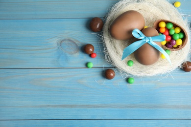 Photo of Flat lay composition with tasty chocolate eggs, colorful candies and decorative nest on light blue wooden table. Space for text