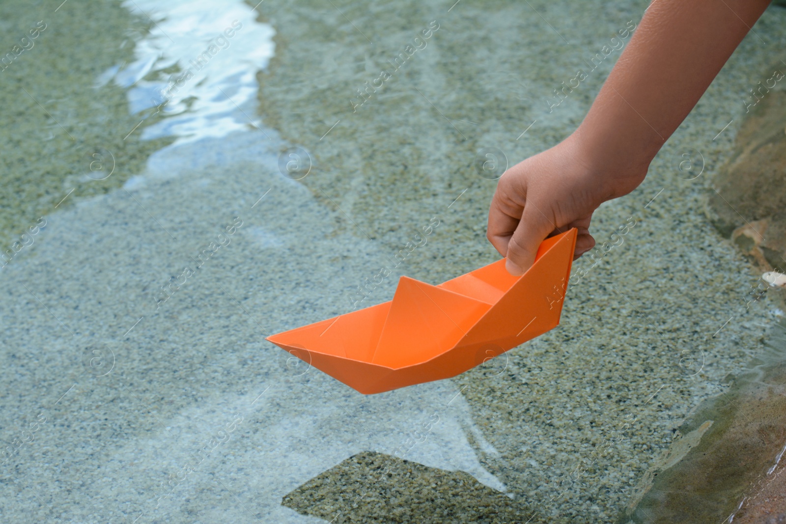 Photo of Kid launching small orange paper boat on water outdoors, closeup