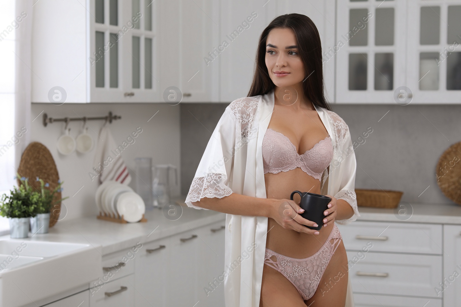 Photo of Young woman with cup wearing elegant underwear and robe in kitchen. Space for text