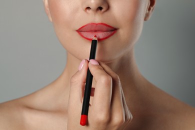 Young woman applying beautiful red lip pencil on grey background, closeup