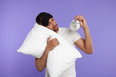 Photo of Tired man with pillow, sleep mask and alarm clock on violet background. Insomnia problem