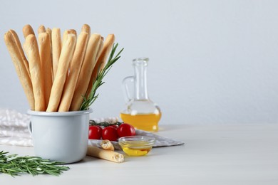 Photo of Delicious grissini sticks, oil, rosemary and tomatoes on white wooden table. Space for text