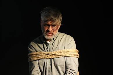 Photo of Beaten man tied with rope on black background. Hostage taking