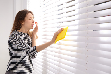 Photo of Young woman suffering from dust allergy while cleaning window blinds at home