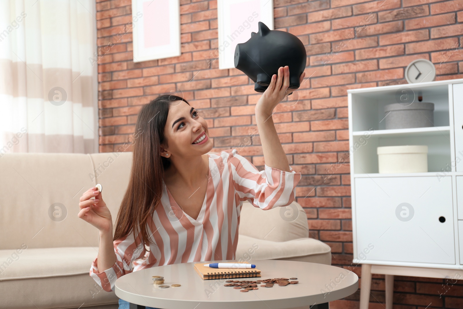 Photo of Happy young woman with piggy bank and money at home