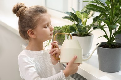 Cute little girl watering beautiful green plant on windowsill at home. House decor