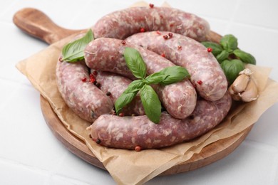 Board with raw homemade sausages, basil leaves and peppercorns on white tiled table, closeup