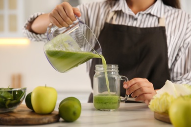 Young woman pouring fresh green juice into mason jar at table in kitchen, closeup