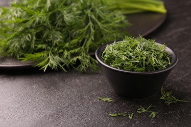 Photo of Sprigs of fresh dill and bowl with cut one on dark textured table, closeup