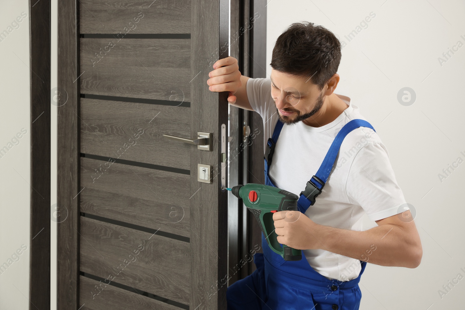 Photo of Worker in uniform with screw gun repairing door lock indoors