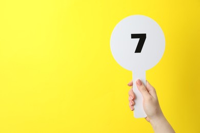 Photo of Woman holding auction paddle with number 7 on yellow background, closeup. Space for text